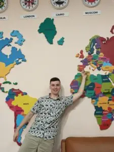 Smiling young man in a tropical-pattern shirt posing in front of a large colorful world map on the wall.
