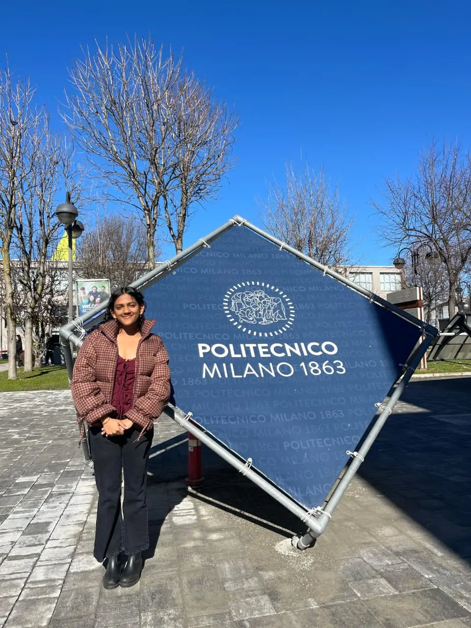 Young woman in a burgundy jacket stands beside a tilted blue sign reading 'Politecnico Milano 1863' outdoors under a clear blue sky.